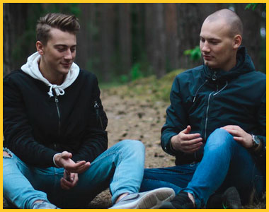 Two young men sitting on the ground in the woods talking